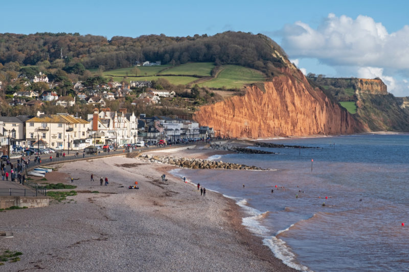 view from high up of the pebble beach at Sidmouth in Devon UK with white painted buildings along the seafront and the red sandstone cliffs of Pennington Point in the background