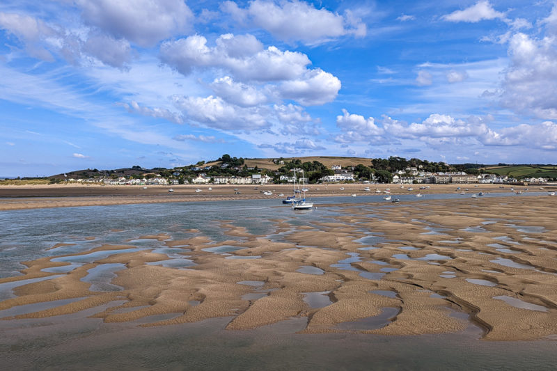 river estuary at low tide with small pools of water between ridges of sand on the river bed, there is a small hill on the far side with a village, and a few small sailing boats on the sandy riverbed