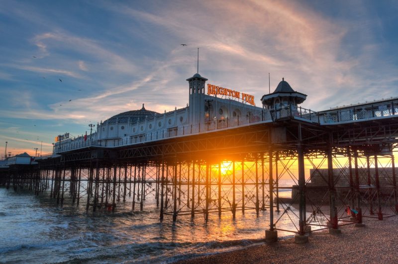 brighton pier shot from low down on th ebeach so the pier is above the horizon, most of the structure is silhouetted against a sunset sky with the setting sun appearing just above the horizon of the sea between the metal poles holding up the pier. best uk cities for nightlife. 