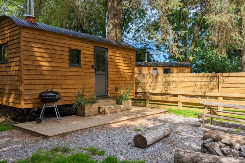 wooden shepherds hut with a grey roof and a small gravel courtyard area in fornt with logs for seats and trees overhead. Glamping essentials packing list. 