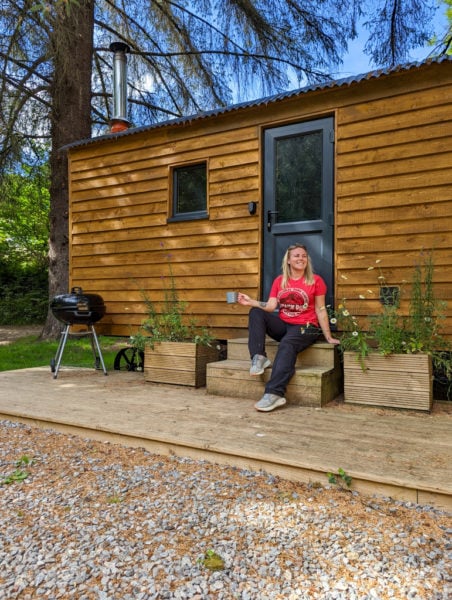 emily wearing black trousers and a bright red t-shirt with the jurassic park logo on it sitting on the wooden steps of a small log cabin next to a pine tree holding a blue mug of coffee. she has long blonde hair worn down and is looking up towards the sky.