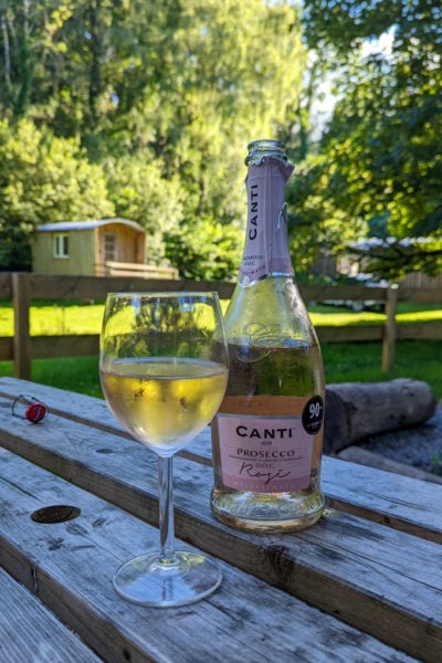 bottle of rose prosecco next to a full wine glass of the same prosecco on a wooden picnic table outside with trees and a small wooden shepherds hut out of focus in the background. Glamping essentials packing list.