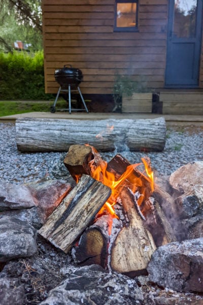 small campfire with logs burning in a stone pit in front of a small wooden shepherds hut with a large pine log for sitting on 
