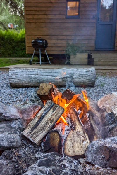 small campfire with logs burning in a stone pit in front of a small wooden shepherds hut with a large pine log for sitting on