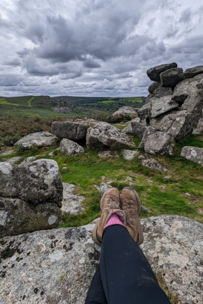 crossed legs in black yoga leggings pink socks and brown hiking legs crossed at the ankles on top of a grey rock in Dartmoor with large grey boulders behind and a view of a grassy countryside landscape beyond underneath a grey cloudy sky