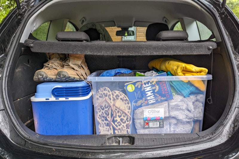 car boot with a large plastic box filled with many items including a book, flip flops, and a sleeping bag, next to a smaller blue coolbox with a pair of brown hiking boots balanced on top