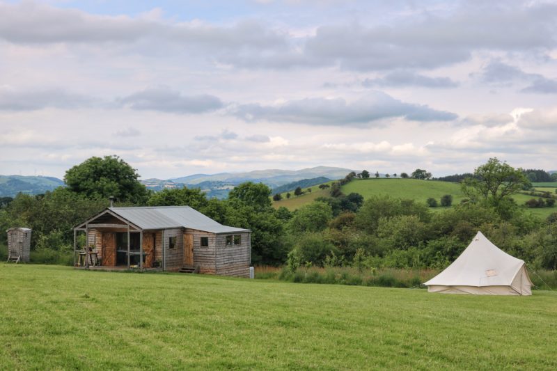 field with a small single storey wooden log cabin on the left and a white canvas bell tent on the right. there are grassy hills behind and cloudy sky overhead - a glamping site in wales