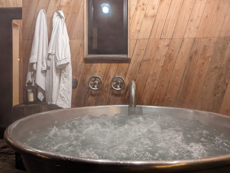grey metal outdoor hot tub with wheel-style taps next to a wooden cabin wall with two white bath robes hanging up