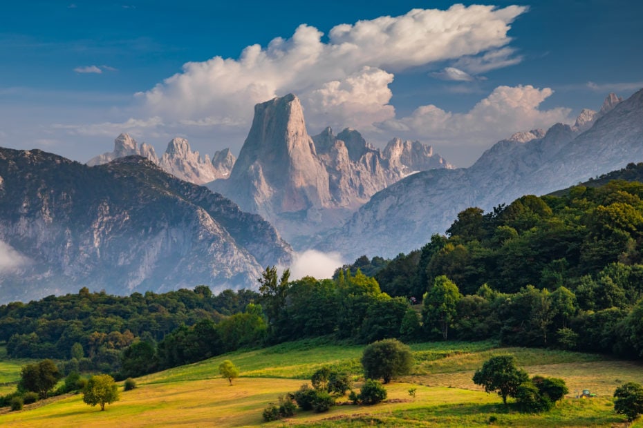 wide valley with woodland and a grassy meadow in front of a mountain scape with a very tall conical mountain peak rising above the others surroudned by clouds. Picos de Europa, best national parks in Spain