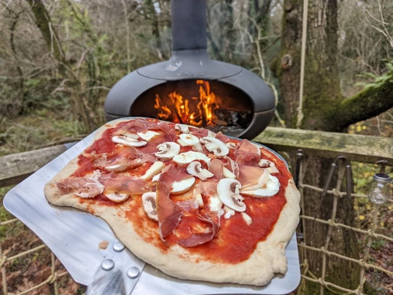 uncooked homemade pizza topped with mushroom slices and ham on a metal shovel being carried towards an outdoor pizza oven at a log cabin glamping site in dorset
