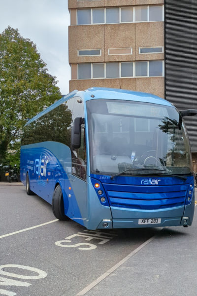 blue RailAir coach in a car park in front of a beige stone office block 