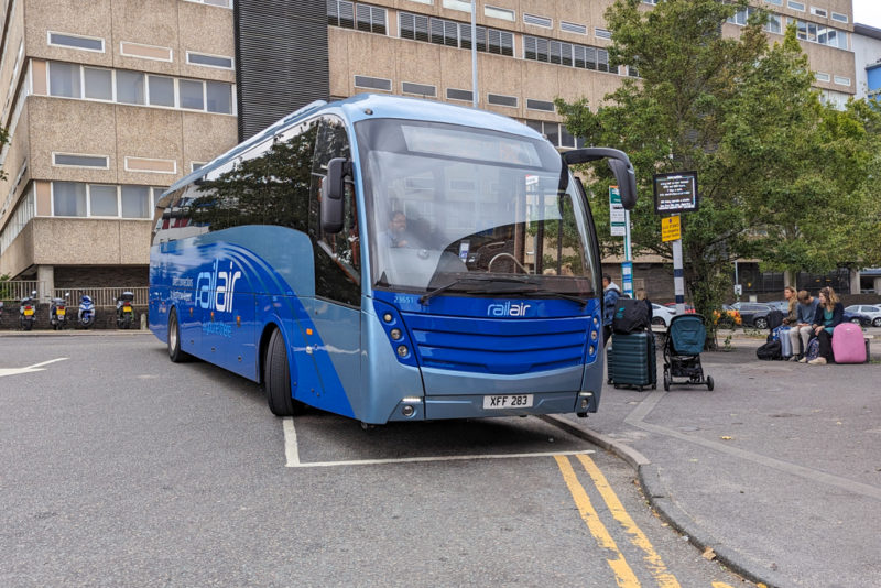 blue RailAir Heathrow bus in Woking station carpark with a beige stone office block in the background