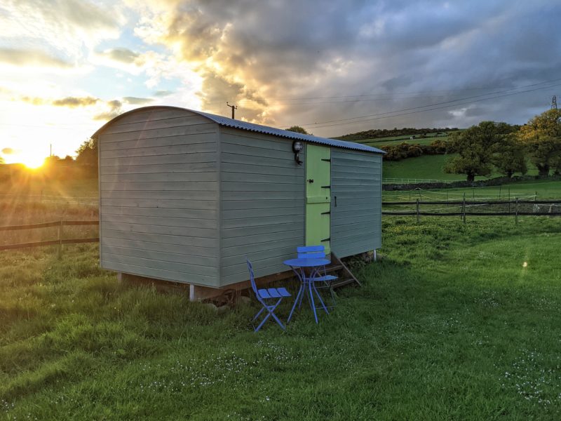 wooden shepherd hut painted light sage green with a yellow door and a grey roof in a field with hills behind and the sun setting just behind the hut