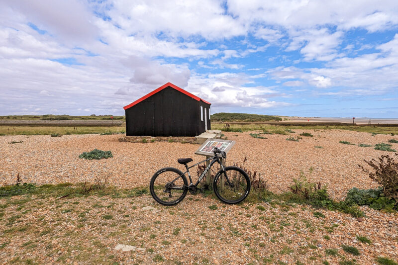 black mountain bike leaning against a sign on a shingle beach with wildflowers growing between the stones and a small black wooden hut with a red roof behind