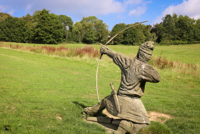 wooden statue of a man in medieval clothes and hat holding a large bow and arrow in a grass field with trees in the distance. Things to do in 1066 Country East Sussex England. 