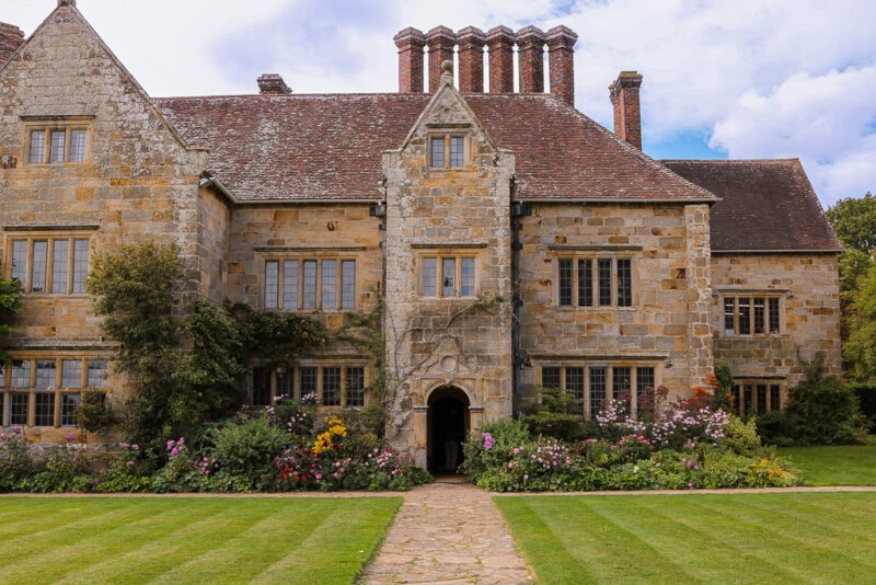 front of a large country manor house built from beige stone with a reddish tiled roof in front of a neat lawn mowed into stripes