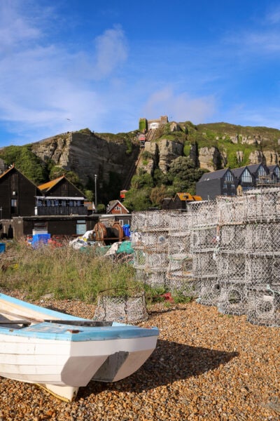 small white rowing boat and a stack of fishing nets on a stoney beach with some buildings behind and a large grassy cliff with a furnicular on it. Things to do in 1066 Country East Sussex England. 