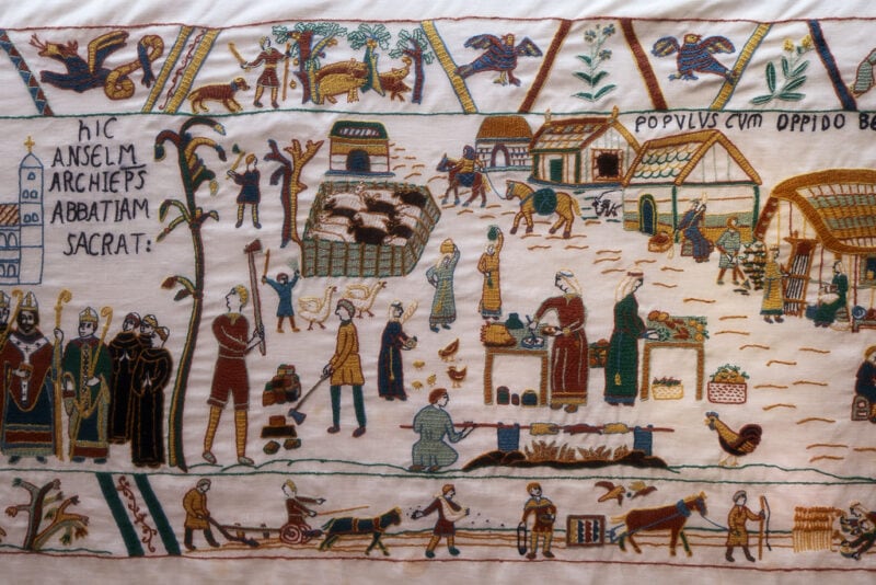 close up of a section of Battle Tapestry, a white cloth emroidered with many figures in muted reds, yellows and greens