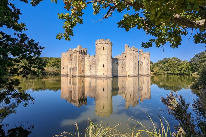 a castle with a moat around it reflected in the still water and framed by trees on a very sunny day