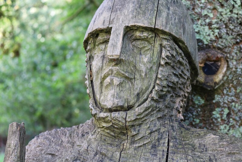 close up of the hed of a wooden sculpture of a man wearing a medieval helmet and armour leaning against a tree