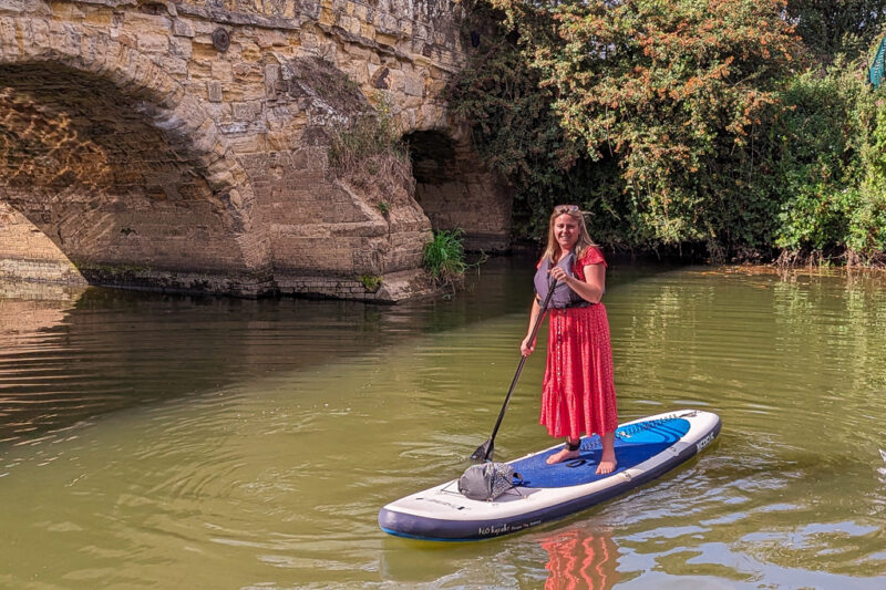 emily wearing a long red dress standing on a paddleboard on a river with a low stone bridge behind. Things to do in 1066 Country East Sussex England. 