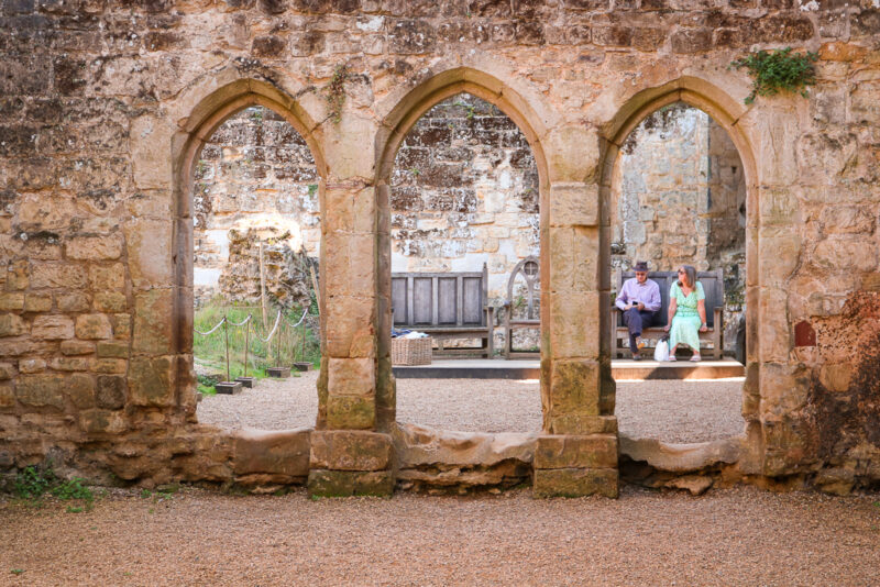 stone wall of a castle ruin with three archways with more of the castle visible behind, through the 3rd arch you can see a man and woman sitting on a wooden bench