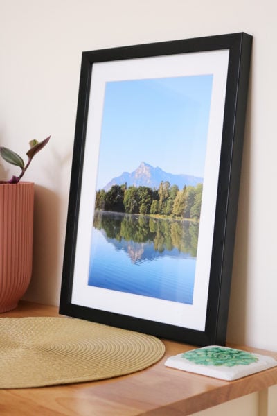 large photo in a blackframe on a wooden table top, the photo shows a mountain reflected in a blue lake