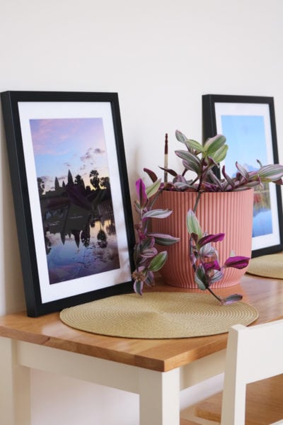 two framed A4 sized photos on a narrow pine wood table with cream chairs against a cream wall with a pink plant pot filled with a purple and green trailing plant in between them. one photo has angkor wat at sunrise and the other has a mountain reflected in a blue lake.