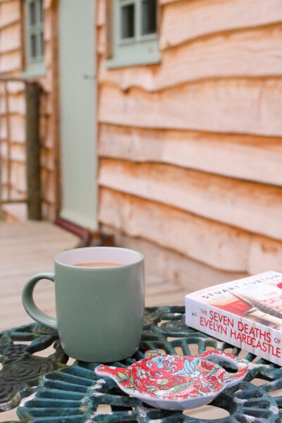 a book and a sage green mug filled with coffee on a green wrought iron table outside a wooden cabin with a sage green front door