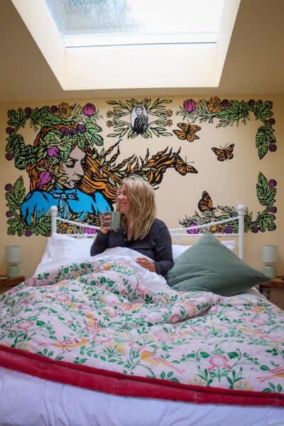 emily wearing a grey long sleeved pyjama top sitting in a bed with pink and yellow patterned cover holding a mug in front of a large painted mural of a woman with long ginger hair.