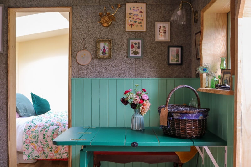 small wooden folding table painted bright green in front of a grey wall with green wooden panels on the lower half. there are many pictures on the wall and a bunch of flowers in a vase next to a wicker basket on the table.