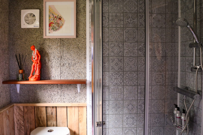 interior of a bathroom in a cabin with grey walls and grey patterned tiles in the shower cubicle. there is a small shelf with an orange statuette of a woman on it.