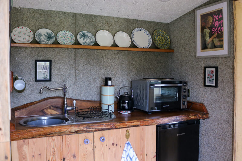 small cabin kitchen with pine wood cupboards with a sink and a microwave, there is a shelf above with several patterned china plates lined up on it. Starcroft Farm Cabins Review