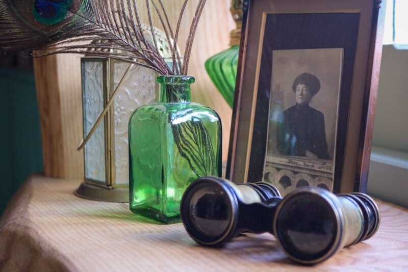 close up of a wooden windowledge with several ornaments on it including some antique brass binoculars and a black and white photo of a Victorian woman in a gold frame. 