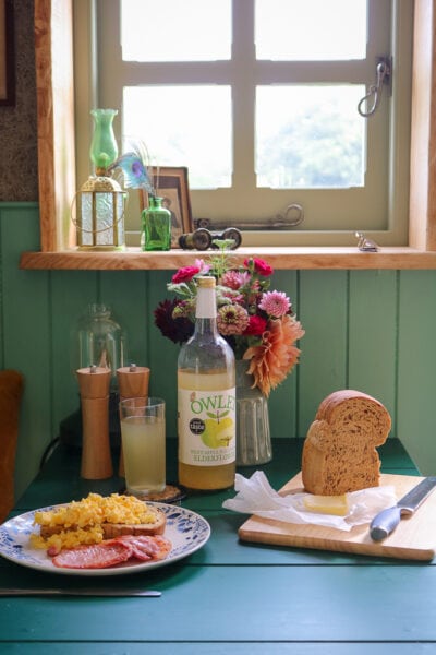 green painted table below a small square window with breakfast laid out, there is a white plate with scrambled eggs and bacon on toast, a loaf of bread on a wooden chopping board, and a large bottle of apple juice