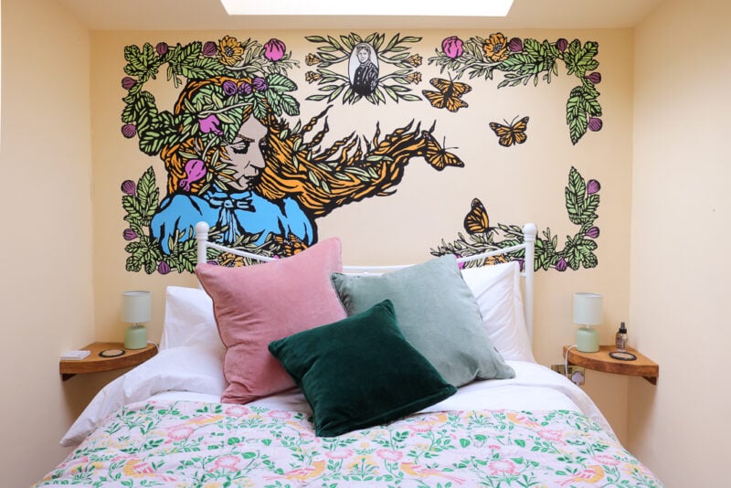 bedroom with yellow walls. There is a double bed with a pink, green and yellow bedspread and green and pink cushions in front of a large full wall mural of a woman with long ginger hair surrounded by leaves, flowers and butterflies. Starcroft Farm Cabins review - glamping Battle, Sussex England. 