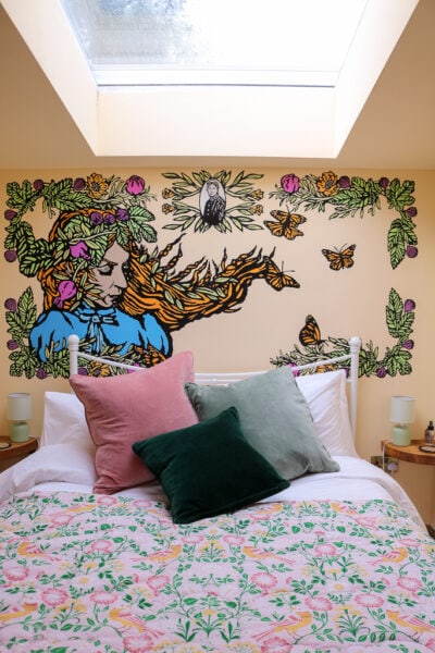bedroom with yellow walls. There is a double bed with a pink, green and yellow bedspread and green and pink cushions in front of a large full wall mural of a woman with long ginger hair surrounded by leaves, flowers and butterflies. Over the bed is a skylight.