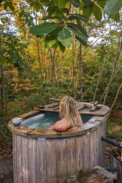 emily in a large wooden hot tub surrounded by woodland, she is facing away from the camera with her elbow on the side of the tub