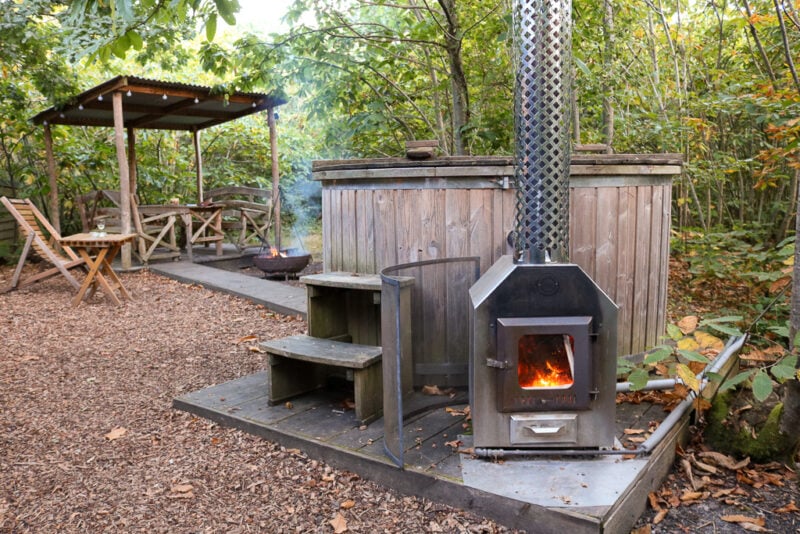 large wooden hot tub with a small log burning stove with metal chimney on the side in a woodland with trees all around an a covered wooden seating area behind with an iron roof. Hot tub at Swallowtail Hill glamping Rye Sussex