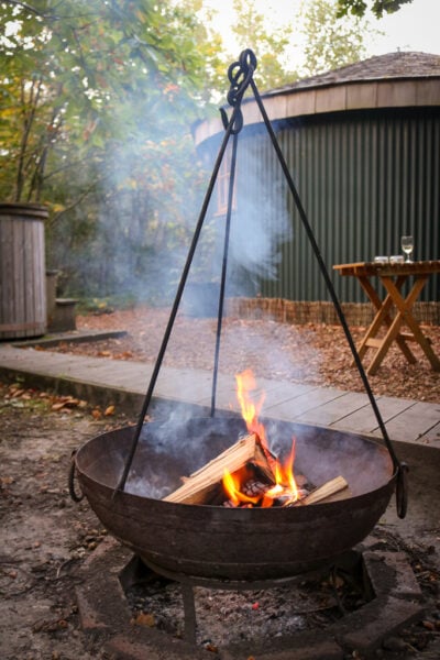 close up of a black cast iron firepit with a small campfire burning in front of a dark green wooden cabin in the woods