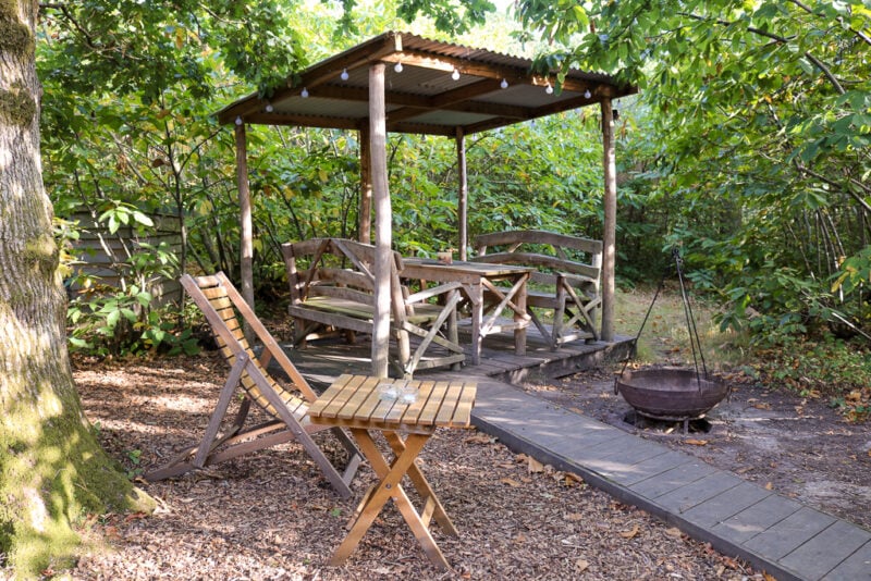 clearing in a woodland with a wooden table and chairs and a cast iron firepit in front of a wooden decking area with an iron roof above a picnic bench