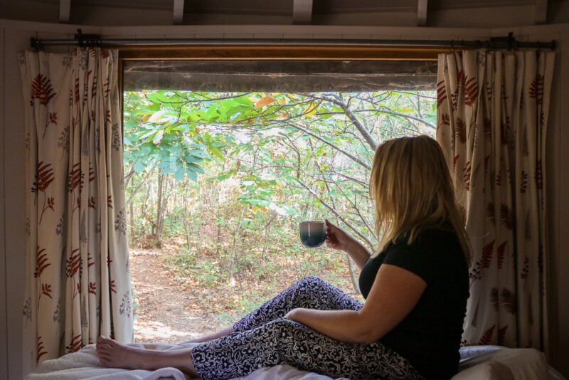 emily sitting at the end of a bed silhouetted against a large window with a view of woodland