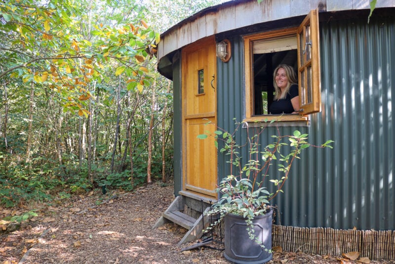circular wooden cabin with green pannelled exterior and a light brown wooden door in a woodland, the window is open and emily is looking out wearing a black t-shirt. Roundhouse Cabin at Swallowtail Hill glamping review. 