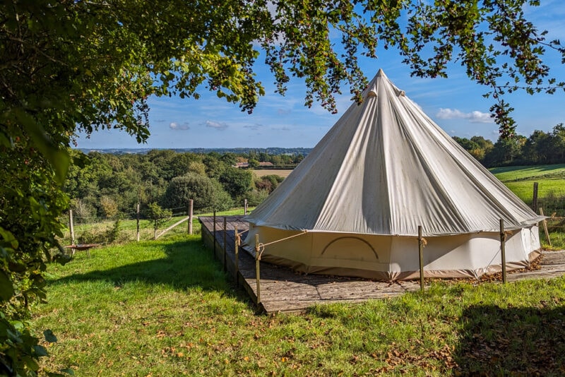 white canvas bell tent in a grass field at the top of a hill with grass sloping down to a dense green woodland in a valley on a sunny day with blue sky above