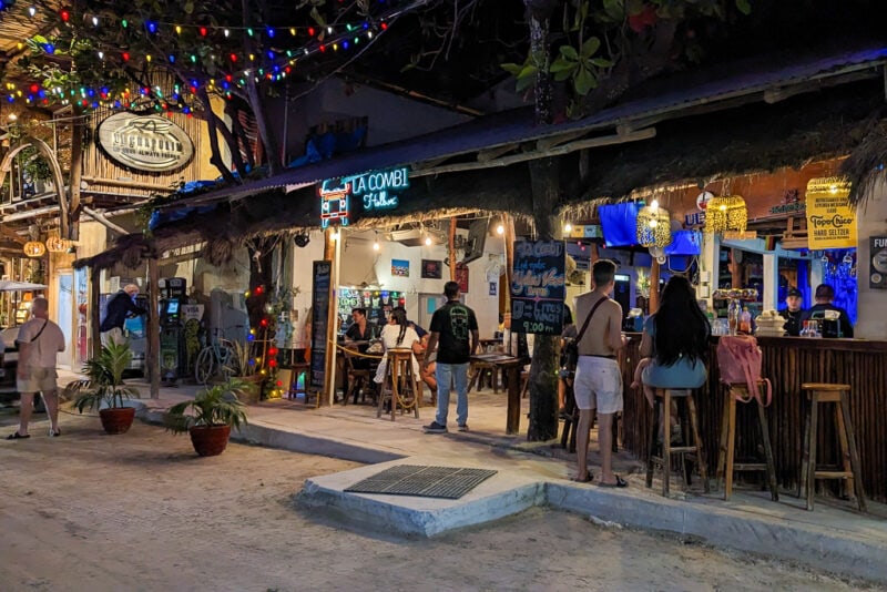 exterior of a crowded open front bar at night on a sandy street with a neon sign with the name La Combi Holbox
