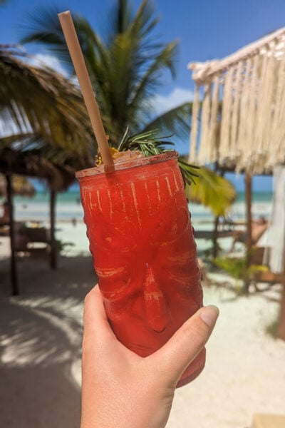 hand holding a tiki style glass with a face carved into it fiulled wiht red cocktail in front of a palm tree with a white sandy bewach and turquoise water behind