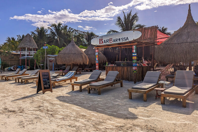 wooden sun loungers with beige cushions in a row on a white sandy beach in front of a wooden bar with a sign that says Barbarossa