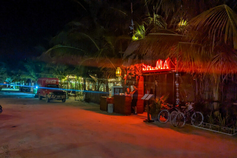 exterior of a bar at night with a grass frond roof and a red neon sign saying "Salma" next to a sandy road