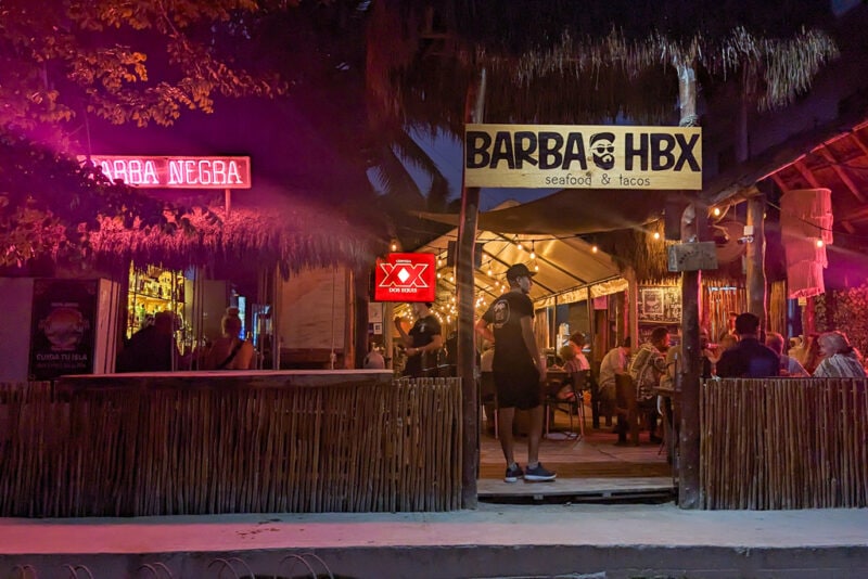 exterior of Barba Negra bar and restaurant in Holbox at night with dried grass roof and a pink neon sign