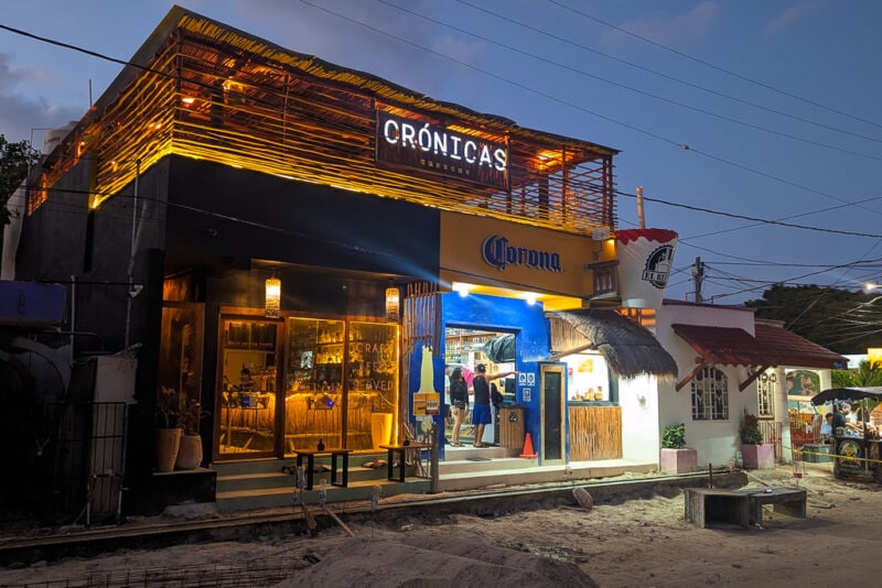exterior of Cronicas taproom lit up at night, a square one storey building with an open front and a roof terrace under a wooden awning